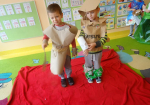 Dwoje dzieci w ekologicznych strojach stoi na czerwonym dywanie.